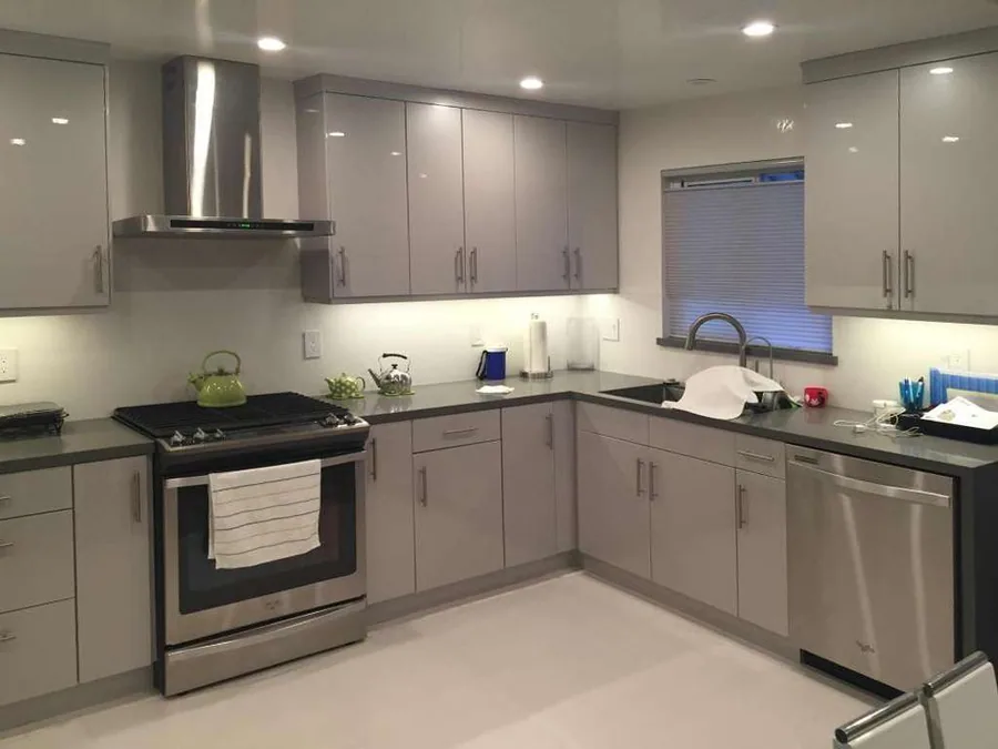Taupe high-gloss flat L-kitchen with chimney hood, stainless chimney hood over a stainless slide-in range, two upper cabinet heights and LED under-cabinet lighting.