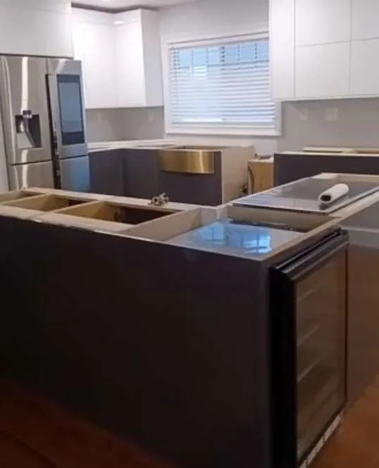 Graphite flat kitchen with white gloss uppers, large L-shaped island, glass-front under-counter wine / beverage refrigerator in the island return and brass-coloured apron sink set in the island.