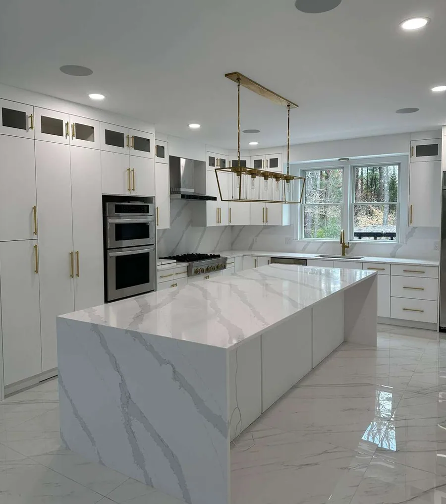 White flat kitchen with brass hardware and a marble waterfall island, full-waterfall island, both ends, frosted / clear glass-front upper cabinets and panel-ready refrigerator column.