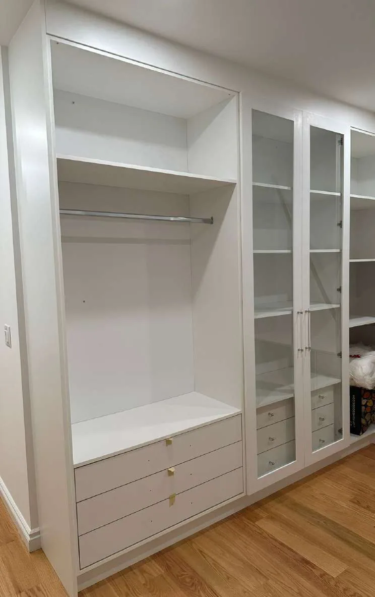 Walk-in closet with bench and glass-front display cabinet, built-in bench seat over a three-drawer bank, open hanging bay with a chrome rail and tall glass-front display cabinet with white interior shelves.