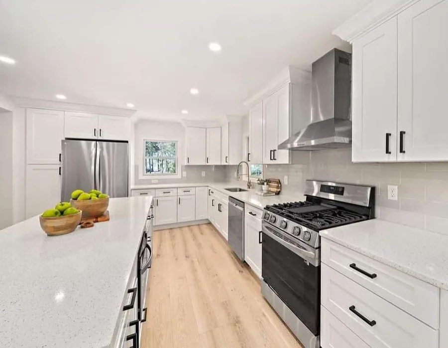 White shaker kitchen with speckled quartz island, matte black bar pulls, stainless chimney hood and stainless gas range with a black glass door.